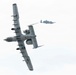 Two military aircraft, identifiable as A-10 Thunderbolt II (Warthog) ground-attack jets, are captured in flight against an overcast sky. The foreground aircraft is performing a steep banking maneuver, revealing its underside with multiple hardpoints and twin rear-mounted turbofan engines. A second identical aircraft is visible in the background at a distance, flying in a more level attitude.