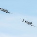 Two A-10 Thunderbolt II (Warthog) aircraft are flying in a diagonal formation against a clear light blue sky. Both aircraft are painted in a gray camouflage scheme and appear to be banking or maneuvering in coordinated flight. The image captures both aircraft from a low-angle perspective, emphasizing their twin-engine rear-mounted configuration and distinctive straight wings.