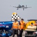 An A-10 Thunderbolt II (Warthog) attack aircraft is captured in flight with landing gear deployed, approaching or departing an airfield during what appears to be an airshow. In the foreground, ground crew personnel wearing orange high-visibility safety vests are visible alongside a blue civilian aerobatic aircraft with a checkered-tail design and a yellow support vehicle. The scene suggests an active airshow environment with multiple aircraft and support staff on the tarmac.