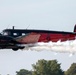 A vintage twin-engine aircraft painted in a striking black and red color scheme performs at what appears to be an airshow, emitting white smoke trails from its fuselage. The aircraft displays markings including 'WAT', a registration number 'N91068', a red star insignia, and references to 'Cleveland'. It is flying at low altitude against an overcast sky with trees visible in the background.