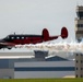 A vintage twin-engine aircraft painted in red and black is performing a low-altitude airshow pass while emitting a dense white smoke trail. The aircraft flies in front of an airport control tower and hangar facilities. The scene is consistent with a military or civilian airshow event.