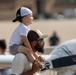 A young child sits on the shoulders of an adult male at what appears to be an airshow or aviation event. Both are wearing baseball caps and looking upward, likely watching aircraft. A small propeller plane is visible in the blurred background.