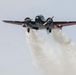 A twin-engine propeller aircraft, dark in color with reddish-brown wing undersides, is photographed from below and slightly in front as it flies overhead. The aircraft is releasing a large white smoke or liquid trail from its underside, consistent with an airshow smoke display or aerial firefighting/retardant drop. The landing gear appears to be extended or partially deployed.