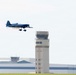 A blue aerobatic aircraft is performing a low-altitude pass over an airfield, trailing smoke from its exhaust. In the background, a prominent air traffic control tower and large aviation hangars are visible. The scene appears to be an airshow or aerobatic demonstration event.