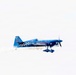 A blue aerobatic propeller aircraft is captured in flight against a pale, overcast sky. The aircraft is emitting a trail of smoke, suggesting it is performing an aerobatic display or airshow maneuver. The plane features distinctive checkered markings and sponsor livery consistent with competitive or exhibition aviation.