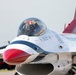 A USAF Thunderbirds F-16 Fighting Falcon is shown on the ground, likely at an airshow. The pilot is visible inside the cockpit wearing a red helmet and raising both fists in a celebratory or acknowledgment gesture. The aircraft displays the distinctive Thunderbirds red, white, and blue livery with stars and stripes markings.