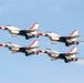 Four U.S. Air Force Thunderbirds aerobatic demonstration team aircraft are flying in a tight diamond formation against a clear blue sky. The jets, identifiable by their distinctive red, white, and blue livery, appear to be performing a precision formation flight maneuver. At least one aircraft is emitting a white smoke trail from its tail.