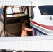The image shows a public aviation event, likely an airshow or open house, where civilians and military personnel are gathered around a small general aviation aircraft. A person in a pink shirt and cap is leaning into the open door of the aircraft, possibly inspecting the cockpit. Military personnel in camouflage uniforms are visible in the background alongside civilian visitors.