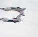 Two United States Air Force Thunderbirds F-16 Fighting Falcons are performing a mirror pass maneuver, with one aircraft flying inverted directly above the other. Both jets display the distinctive red, white, and blue Thunderbirds livery with visible aircraft numbers (5 and 6). Smoke trails are visible behind both aircraft against an overcast sky.