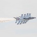 The image shows a tight formation of USAF Thunderbirds aerobatic demonstration team aircraft, identifiable by their distinctive red, white, and blue livery. Four F-16 Fighting Falcon jets are flying in close formation, leaving a white smoke trail behind them. The aircraft appear to be performing a formation maneuver at an airshow.
