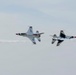 Two U.S. Air Force Thunderbirds demonstration jets perform a close opposing pass maneuver against an overcast sky. Both aircraft are F-16 Fighting Falcons painted in the distinctive red, white, and blue Thunderbirds livery. One aircraft is flying upright while the other appears inverted, with white smoke trails visible behind them.