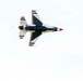 A U.S. Air Force Thunderbirds F-16 Fighting Falcon is captured in flight from a below-and-behind perspective, showing the aircraft's underside. The jet displays the iconic red, white, and blue Thunderbirds livery with the distinctive delta wing configuration. The aircraft appears to be performing an aerobatic maneuver at an air show.
