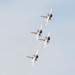 Four military jet aircraft are flying in a tight diagonal formation against a pale gray-blue sky. The jets display a distinctive red, white, and blue color scheme consistent with the U.S. Air Force Thunderbirds demonstration team. The aircraft appear to be performing a precision aerobatic maneuver, viewed from below at an angle.