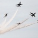 Six military jet aircraft, identifiable as USAF Thunderbirds based on their distinctive white, red, and blue livery, are performing an aerobatic maneuver in an overcast sky. The jets appear to be executing a burst or breakaway formation, diverging outward from a central point with white smoke trails streaming behind them. This is consistent with a precision aerial demonstration team performance at an airshow.
