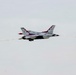 A U.S. Air Force Thunderbirds F-16 Fighting Falcon is captured in flight against an overcast sky. The aircraft displays the distinctive red, white, and blue Thunderbirds livery and is producing a white smoke trail behind it. This appears to be an airshow performance maneuver.