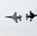 Two U.S. Air Force Thunderbirds F-16 Fighting Falcon jets are performing an aerial demonstration maneuver, appearing to pass in close proximity to each other. One aircraft is flying right-side up while the other is inverted, a classic Thunderbirds opposing pass maneuver. Both aircraft display the distinctive red, white, and blue Thunderbirds livery.