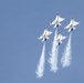 Four USAF Thunderbirds aerobatic demonstration jets are flying in a tight diamond formation against a clear blue sky. The aircraft are white with red and blue markings and are producing white smoke trails from their engines. The formation appears to be climbing or maneuvering during an airshow performance.