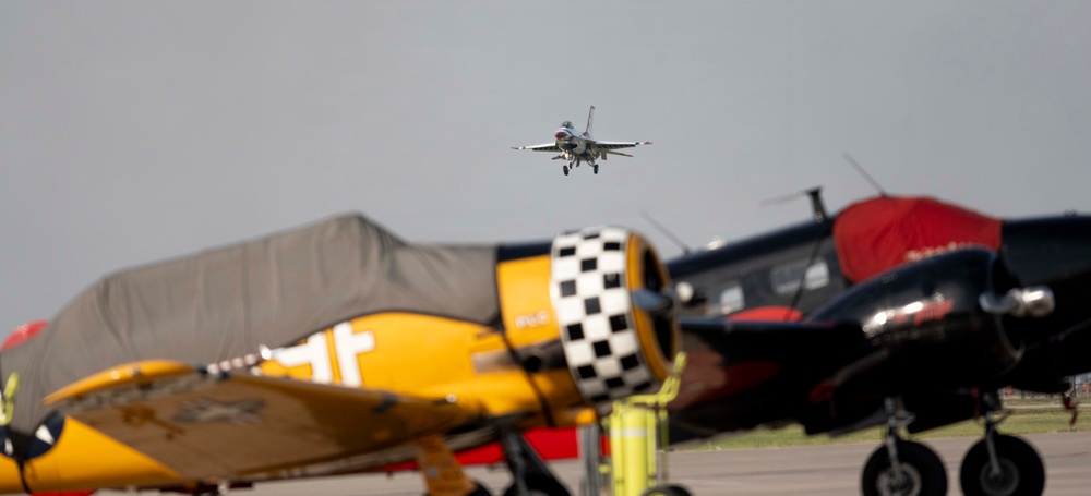 An airshow scene featuring a modern jet fighter aircraft flying in the background while vintage/warbird aircraft are parked on the tarmac in the foreground. The foreground aircraft include a yellow trainer-style plane with a black-and-white checkered engine cowling and a dark-colored aircraft with red markings. The depth of field creates a sharp contrast between the blurred foreground aircraft and the in-flight jet.