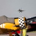 An airshow scene featuring a modern jet fighter aircraft flying in the background while vintage/warbird aircraft are parked on the tarmac in the foreground. The foreground aircraft include a yellow trainer-style plane with a black-and-white checkered engine cowling and a dark-colored aircraft with red markings. The depth of field creates a sharp contrast between the blurred foreground aircraft and the in-flight jet.