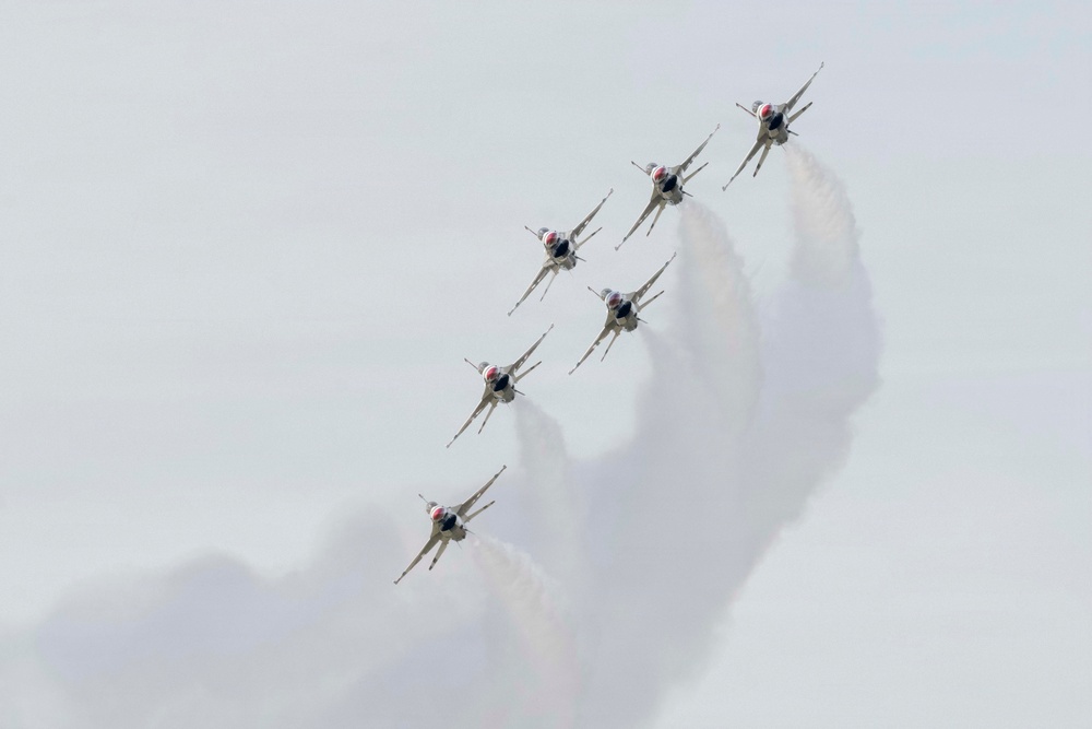 Six military jet aircraft are flying in a tight diagonal echelon formation, trailing white smoke against an overcast gray sky. The aircraft appear to be the U.S. Air Force Thunderbirds aerobatic demonstration team, identifiable by their distinctive red, white, and blue livery. The formation is climbing or banking at an angle, producing a dramatic smoke trail effect.