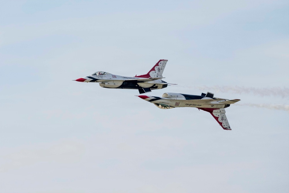 Two U.S. Air Force Thunderbirds F-16 Fighting Falcon jets are performing a close formation maneuver during an airshow. The lower aircraft is flying inverted (upside down) while the upper aircraft flies in normal orientation, demonstrating a signature Thunderbirds pass. Both aircraft display the distinctive red, white, and blue Thunderbirds livery.