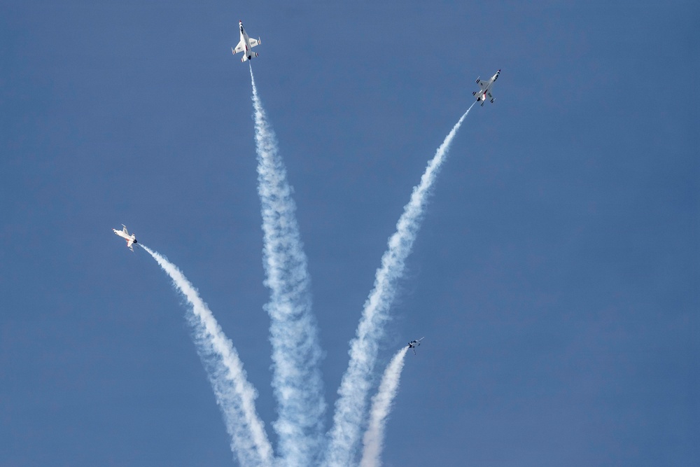 Four military jet aircraft are performing an aerobatic maneuver, diverging outward from a central point in a burst or 'bomb burst' pattern against a clear blue sky. Each aircraft is trailing a white smoke contrail, creating a symmetrical fan-like visual effect. The formation suggests a precision aerobatic team demonstration, consistent with groups like the U.S. Air Force Thunderbirds.