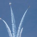 Four military jet aircraft are performing an aerobatic maneuver, diverging outward from a central point in a burst or 'bomb burst' pattern against a clear blue sky. Each aircraft is trailing a white smoke contrail, creating a symmetrical fan-like visual effect. The formation suggests a precision aerobatic team demonstration, consistent with groups like the U.S. Air Force Thunderbirds.