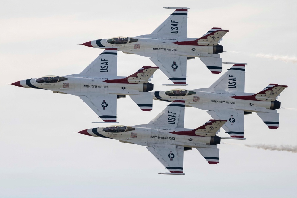 Four U.S. Air Force Thunderbirds aerobatic demonstration team aircraft are flying in a tight diamond formation. The jets, identified as F-16 Fighting Falcons, display the distinctive red, white, and blue Thunderbirds livery with 'USAF' and 'United States Air Force' markings. At least one aircraft is producing a visible vapor/smoke trail from its engine exhaust.