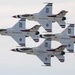 Four U.S. Air Force Thunderbirds aerobatic demonstration team aircraft are flying in a tight diamond formation. The jets, identified as F-16 Fighting Falcons, display the distinctive red, white, and blue Thunderbirds livery with 'USAF' and 'United States Air Force' markings. At least one aircraft is producing a visible vapor/smoke trail from its engine exhaust.