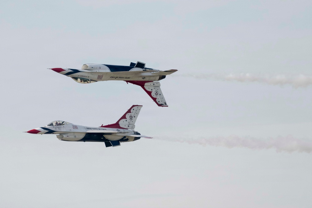 Two U.S. Air Force Thunderbirds F-16 Fighting Falcon jets are performing an aerobatic maneuver at an airshow. One aircraft is flying inverted (upside down) while the other flies in normal orientation, passing in close proximity. Both jets are trailing white smoke and display the distinctive red, white, and blue Thunderbirds livery.