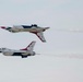 Two U.S. Air Force Thunderbirds F-16 Fighting Falcon jets are performing an aerobatic maneuver at an airshow. One aircraft is flying inverted (upside down) while the other flies in normal orientation, passing in close proximity. Both jets are trailing white smoke and display the distinctive red, white, and blue Thunderbirds livery.