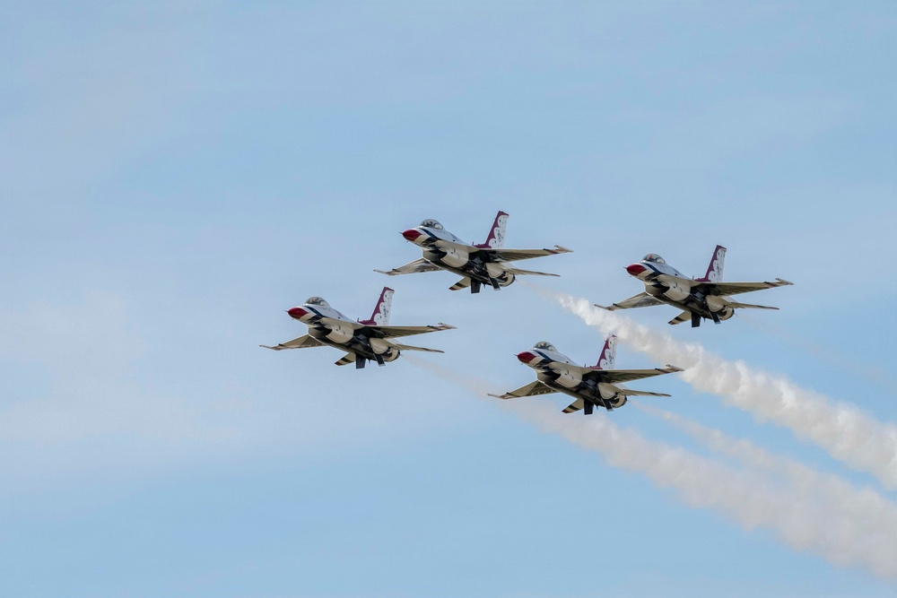 Four U.S. Air Force Thunderbirds demonstration team aircraft fly in a tight diamond formation against a clear blue sky. The jets, identifiable by their distinctive red, white, and blue livery, are producing white smoke trails from at least two of the aircraft. This appears to be an airshow performance showcasing precision formation flying.