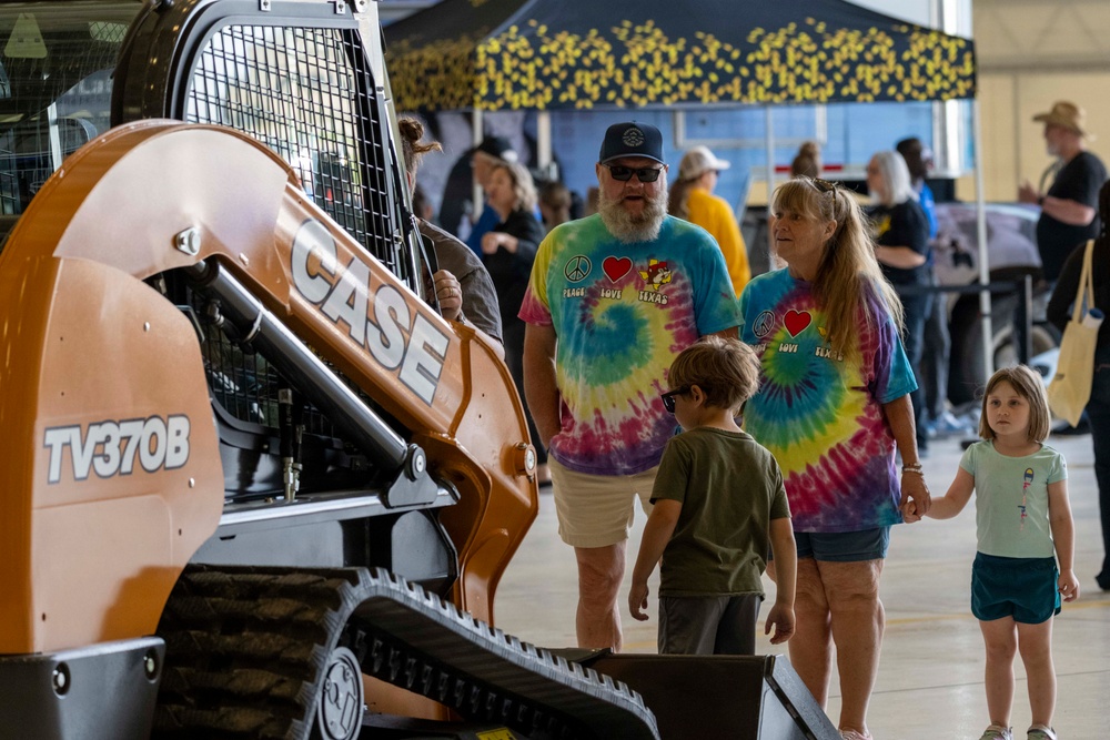 A family group is viewing a Case TV370B compact track loader at what appears to be an indoor equipment exhibition or trade show. Two adults wearing matching tie-dye 'Peace Love Texas' t-shirts are accompanied by two young children examining the orange construction machine. The event is held in a large indoor venue with additional exhibitor tents and visitors visible in the background.