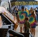 A family group is viewing a Case TV370B compact track loader at what appears to be an indoor equipment exhibition or trade show. Two adults wearing matching tie-dye 'Peace Love Texas' t-shirts are accompanied by two young children examining the orange construction machine. The event is held in a large indoor venue with additional exhibitor tents and visitors visible in the background.