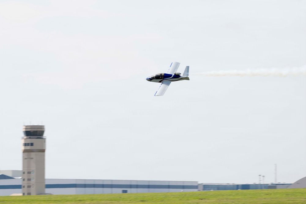 A small aerobatic aircraft with blue and white livery is performing a low-altitude pass over an airport, trailing white smoke from its tail. The aircraft appears to be in a slight bank, suggesting an airshow or aerobatic demonstration. An air traffic control tower and large hangar building are visible in the background.