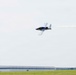 A small aerobatic aircraft with blue and white livery is performing a low-altitude pass over an airport, trailing white smoke from its tail. The aircraft appears to be in a slight bank, suggesting an airshow or aerobatic demonstration. An air traffic control tower and large hangar building are visible in the background.