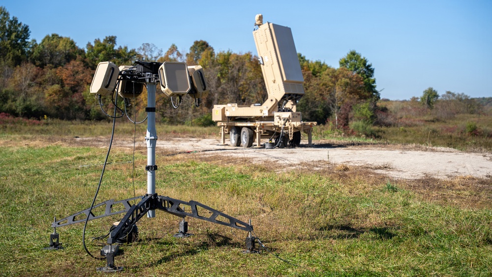 The image shows a military field setup featuring two distinct pieces of equipment in an open grassy area. In the foreground is a small tripod-mounted sensor or antenna array with multiple panels and cabling, while in the background sits a large tan/desert-colored trailer-mounted radar or antenna system in a raised/deployed position. The setting appears to be a military test or demonstration site with tree lines visible in the background under clear blue skies.