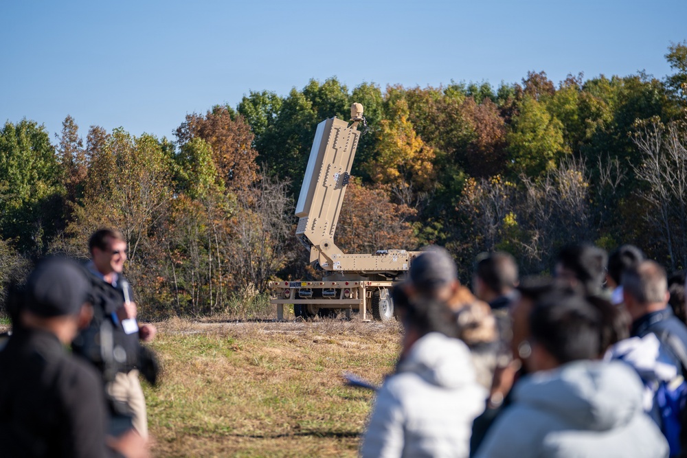 A group of observers watches a military missile launcher or radar system mounted on a flatbed trailer in an open field. The system appears to be in an elevated/deployed position, suggesting a demonstration or test event. The autumn foliage in the background and the civilian-appearing crowd suggest this may be a defense industry demonstration or media event.