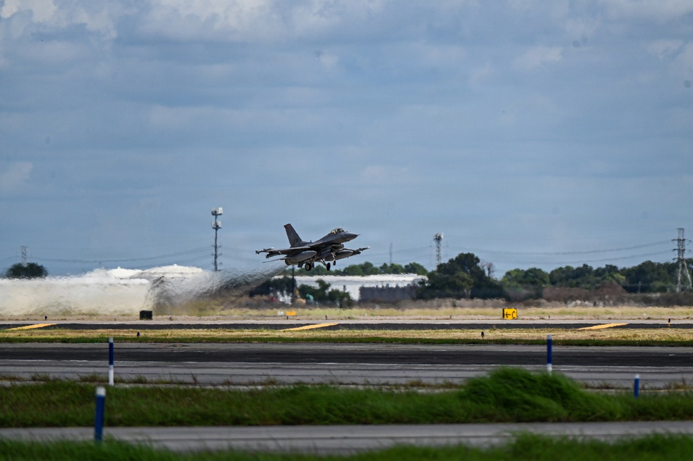 A military fighter jet, consistent with an F-16 Fighting Falcon configuration, is captured at the moment of takeoff from a paved runway. The aircraft's landing gear is still extended as it lifts off, with jet exhaust and spray visible behind it. The setting appears to be a military airfield with surrounding vegetation, utility towers, and overcast skies.